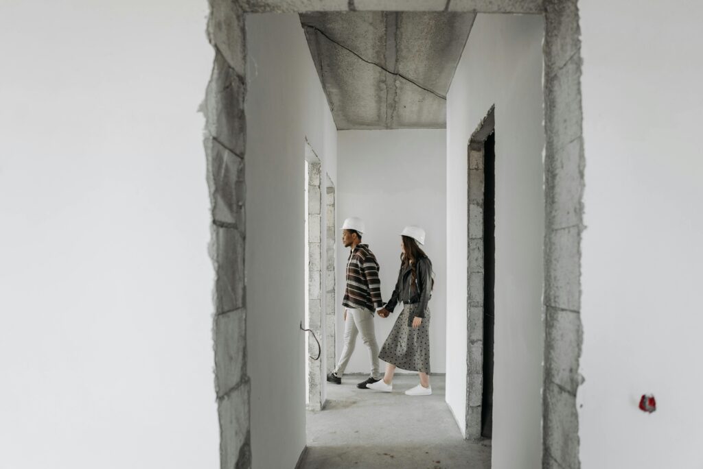A couple holding hands and exploring an unfinished house while wearing hard hats.