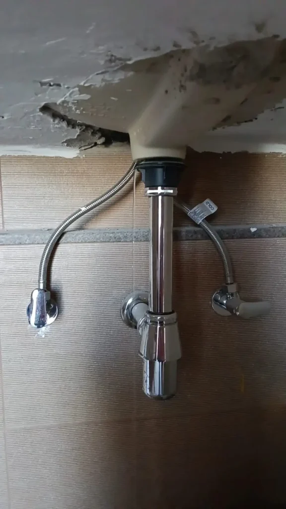 Water leakage beneath bathroom sink with damaged ceiling surface and faulty plumbing connections in a residential property.