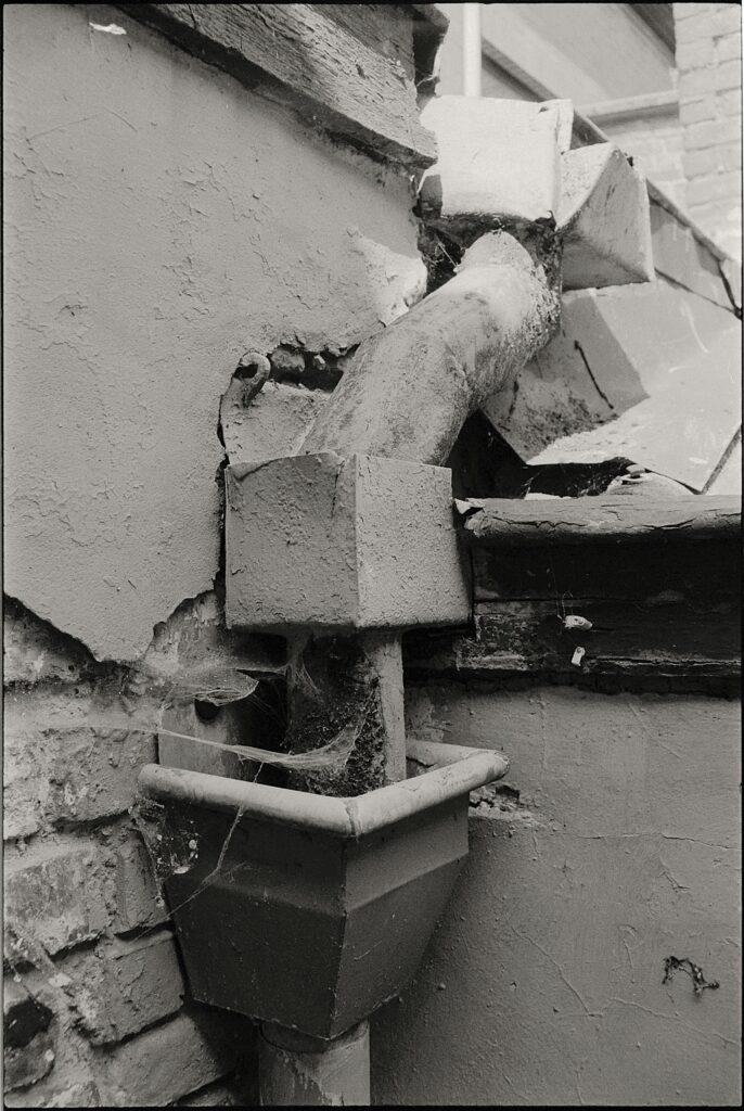 Pexels-photo-16934775-16934775 Close-up of a rusty drain pipe on a deteriorating building wall with visible damage.