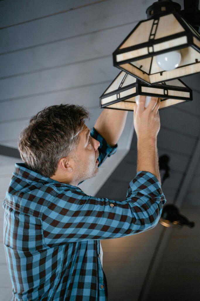 Adult male fixing pendant light indoors, wearing plaid long sleeves. Home maintenance scene.